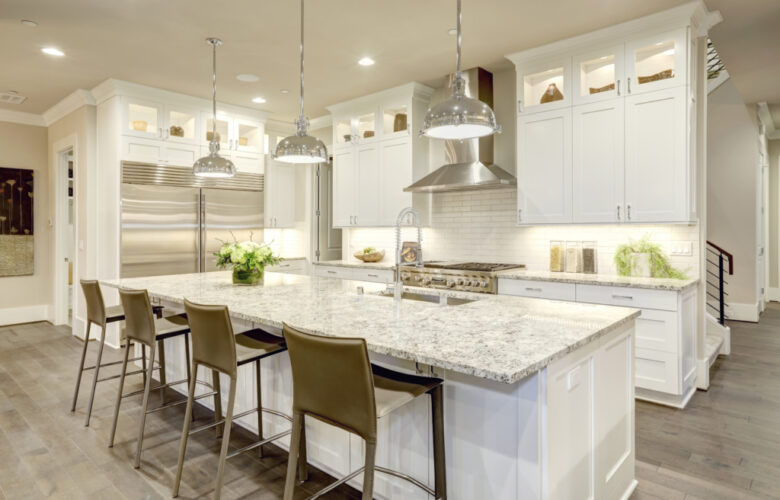 White kitchen design features large bar style kitchen island with granite countertop illuminated by modern pendant lights. Stainless steel appliances framed by white shaker cabinets . Northwest, USA